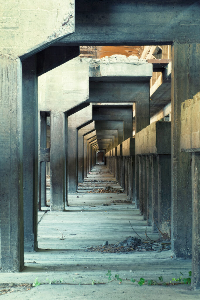 Perspective de portiques et de poutres en béton dans une ancienne usine d’aluminium italienne, éclairée par une ouverture en toiture.