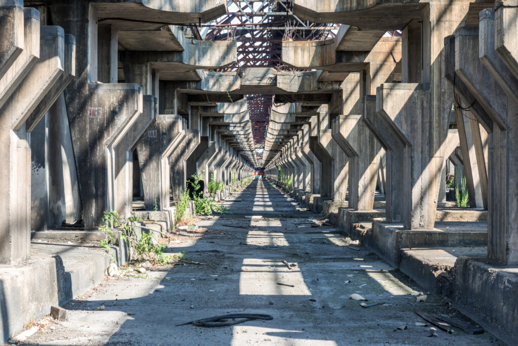 Longue nef industrielle de la SAVA Alumix à Fusina, bordée de piliers en béton et traversée par la lumière et les premières plantes.