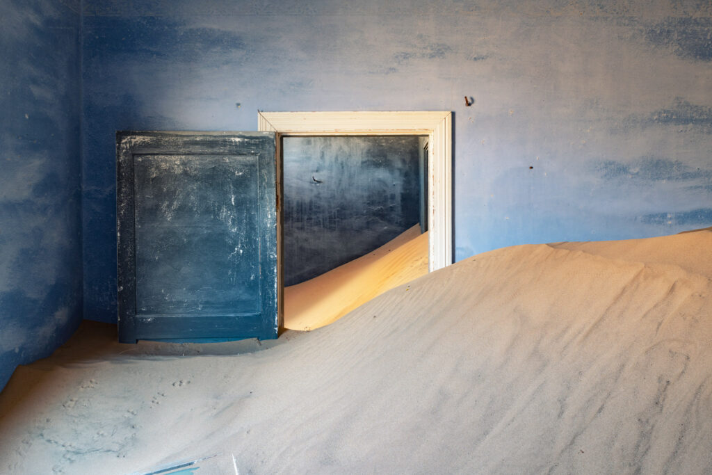 Pièce abandonnée envahie par le sable à Kolmanskop, avec porte bleue, lumière naturelle et traces animales au sol.