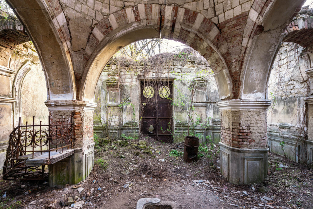 Intérieur ruiné du Beit Kaddishim du cimetière juif de Chișinău, sous des arches de pierre, face à une porte métallique marquée d’impacts