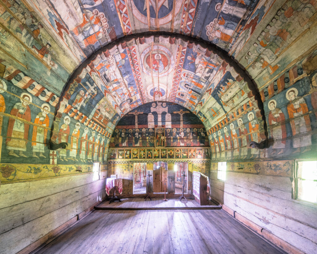 Intérieur d’une église traditionnelle en bois roumaine, avec iconostase, voûte peinte, scènes religieuses polychromes et mobilier liturgique en bois.