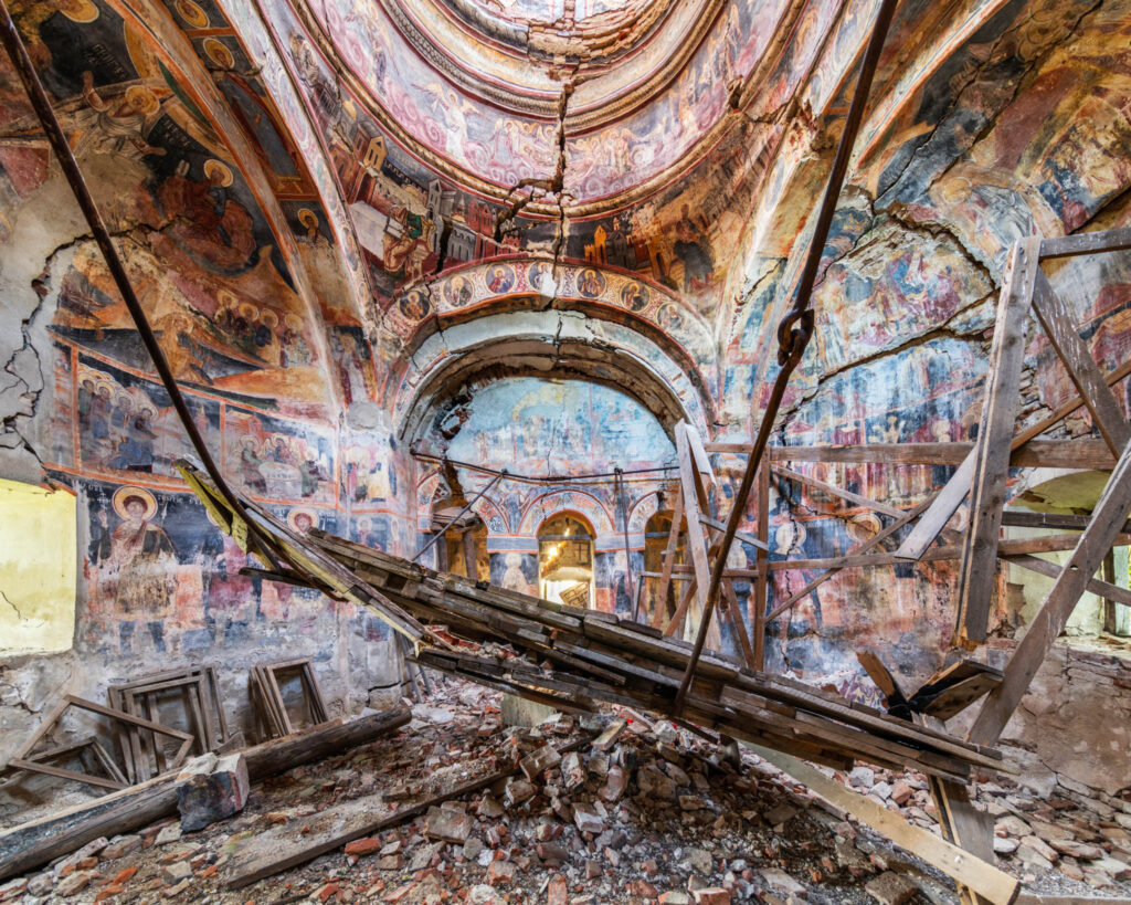 Intérieur d’une grande église orthodoxe rurale roumaine, avec fresques anciennes, tribunes en bois et peintures murales dégradées.
