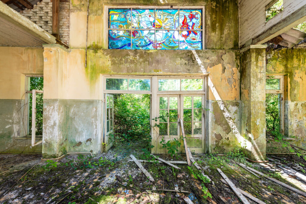 Réfectoire abandonné d’un ancien camp de pionniers soviétique, envahi par la végétation, avec un vitrail presque intact