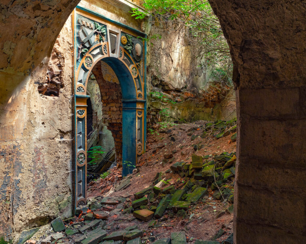 Porte cintrée peinte en bleu, ocre et noir demeurant debout parmi les briques, les enduits dégradés et la végétation d’un palais polonais en ruine