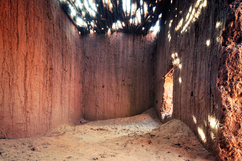 Intérieur d’un ancien poste de douane en terre crue dans le Sahara marocain, avec murs ocre, sol sableux et lumière traversant une toiture de branchages