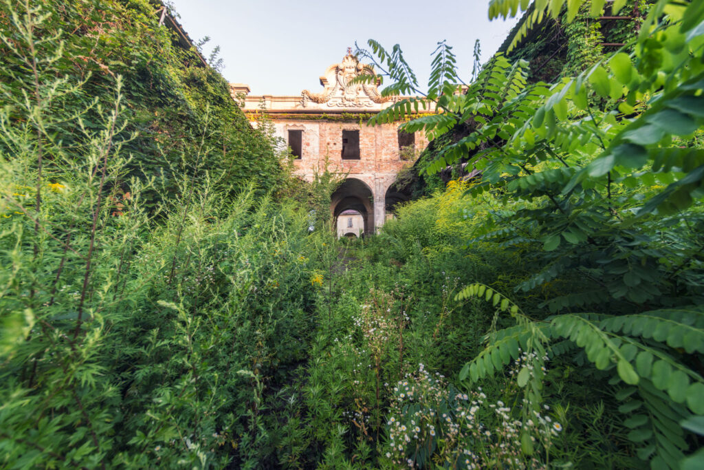 Façade d’une villa historique lombarde en ruine, avec portail central et architecture des XVIIe et XVIIIe siècles dissimulée par une végétation dense