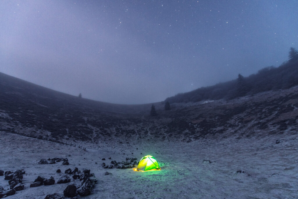 Tente éclairée de nuit dans un paysage enneigé du Puy Pariou, sous un ciel étoilé, Massif central, hiver 2021.