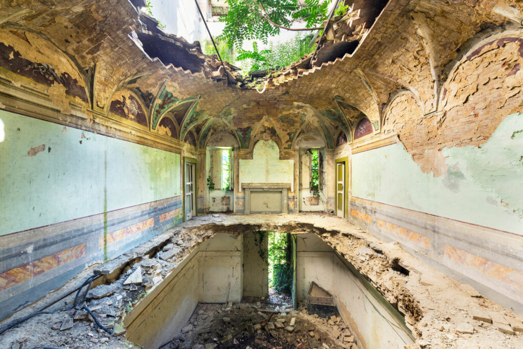 Salle décorée d’un palais italien abandonné, avec fresques murales, plafond effondré, sol ouvert et végétation entrant par les ouvertures.